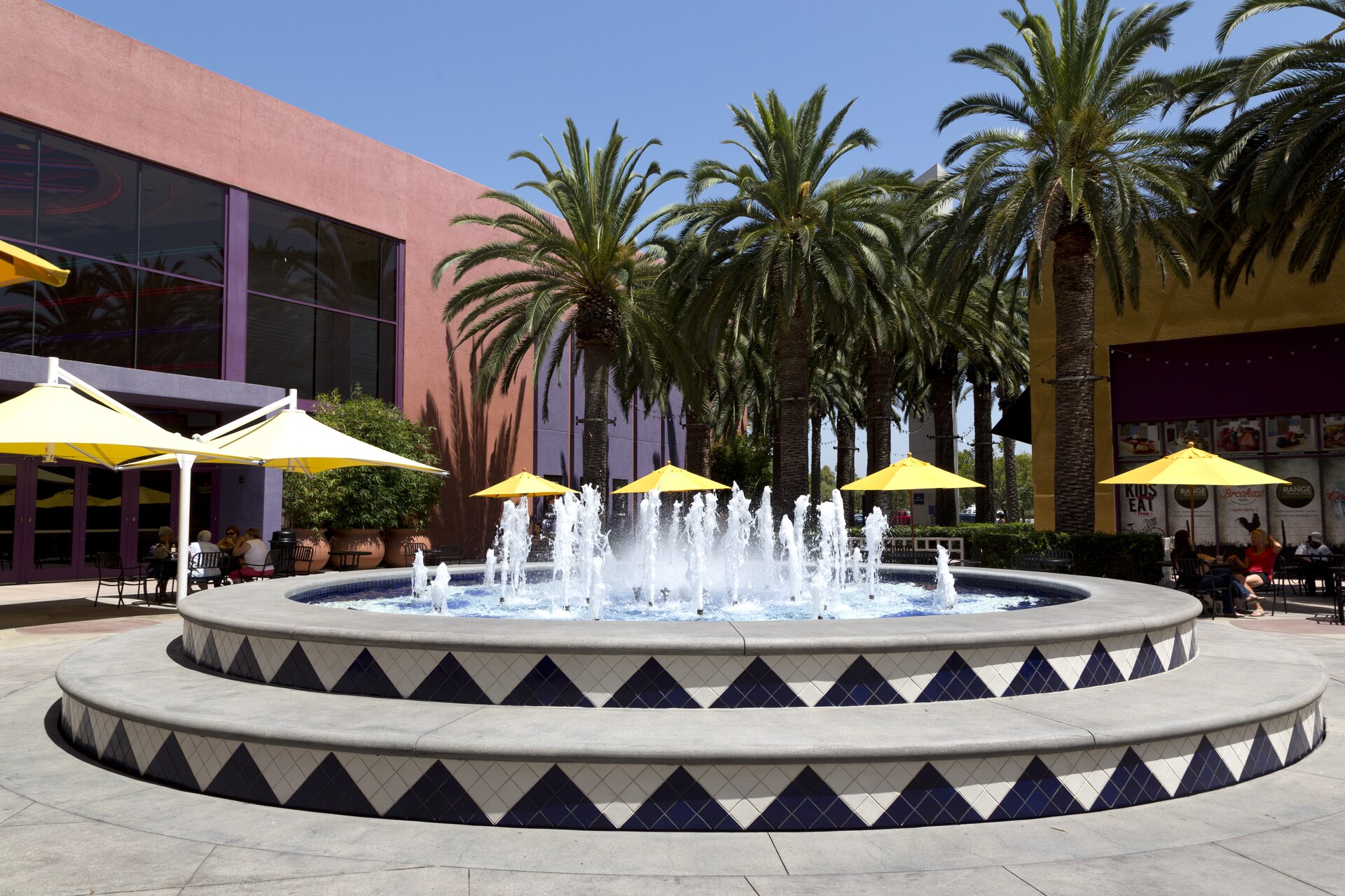 Tiled circular fountain running in an open-air Irvine shopping center plaza, yellow cafe umbrellas and date palms against stucco storefronts