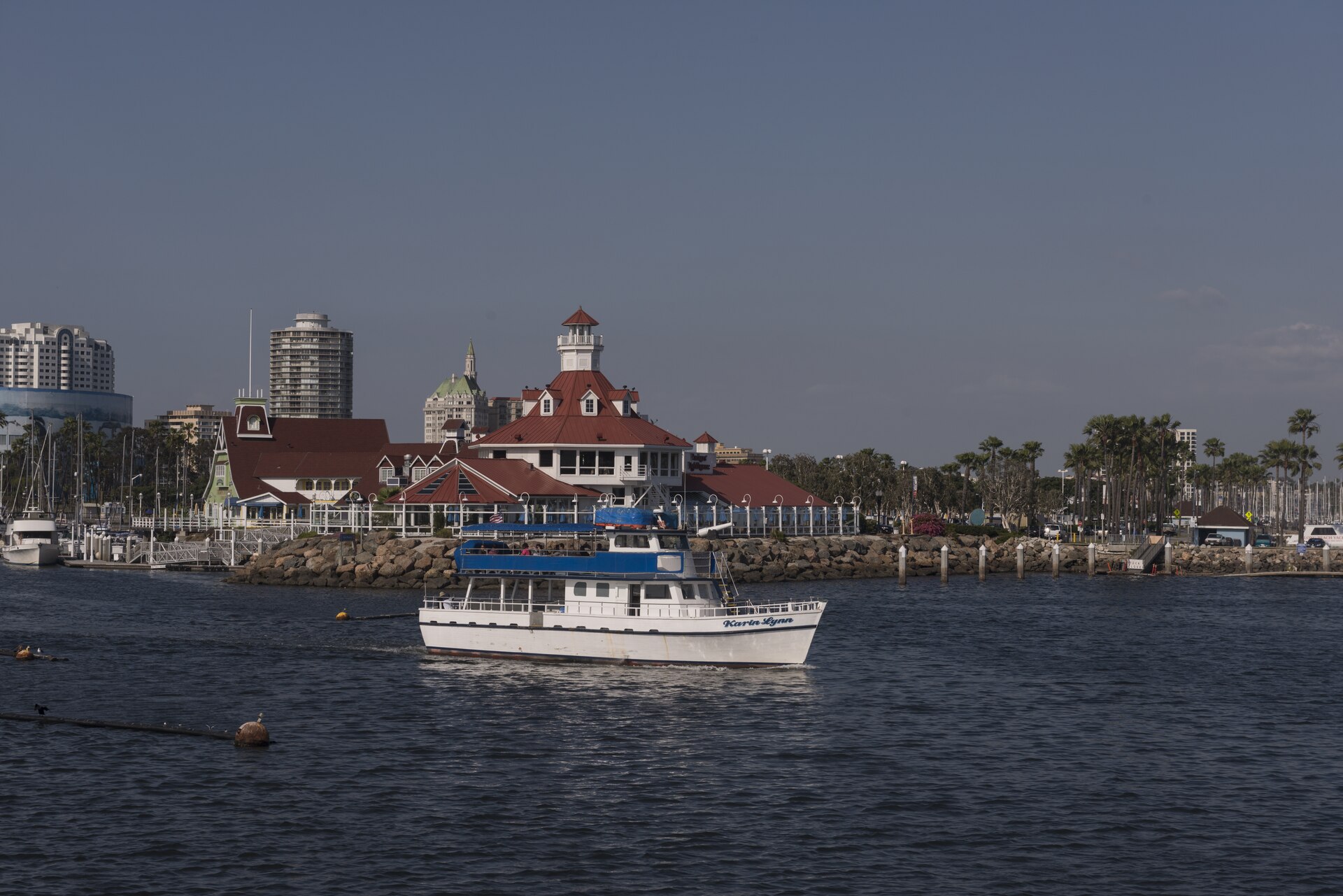 A white passenger boat crossing Long Beach harbor past red-roofed waterfront buildings, a rock breakwater, marina docks and downtown towers