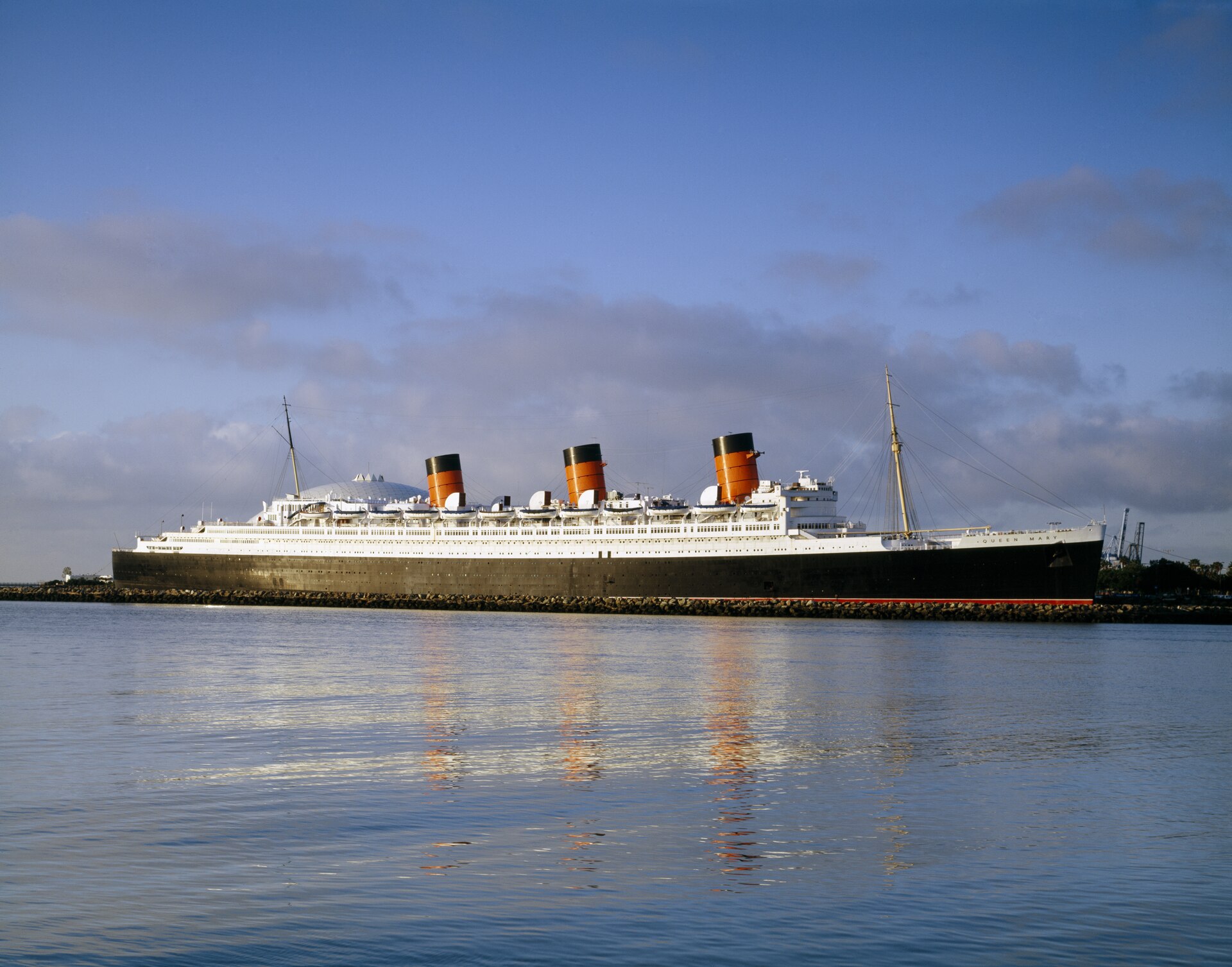 The ocean liner Queen Mary moored at her Long Beach berth, black hull and three orange funnels reflected in flat harbor water