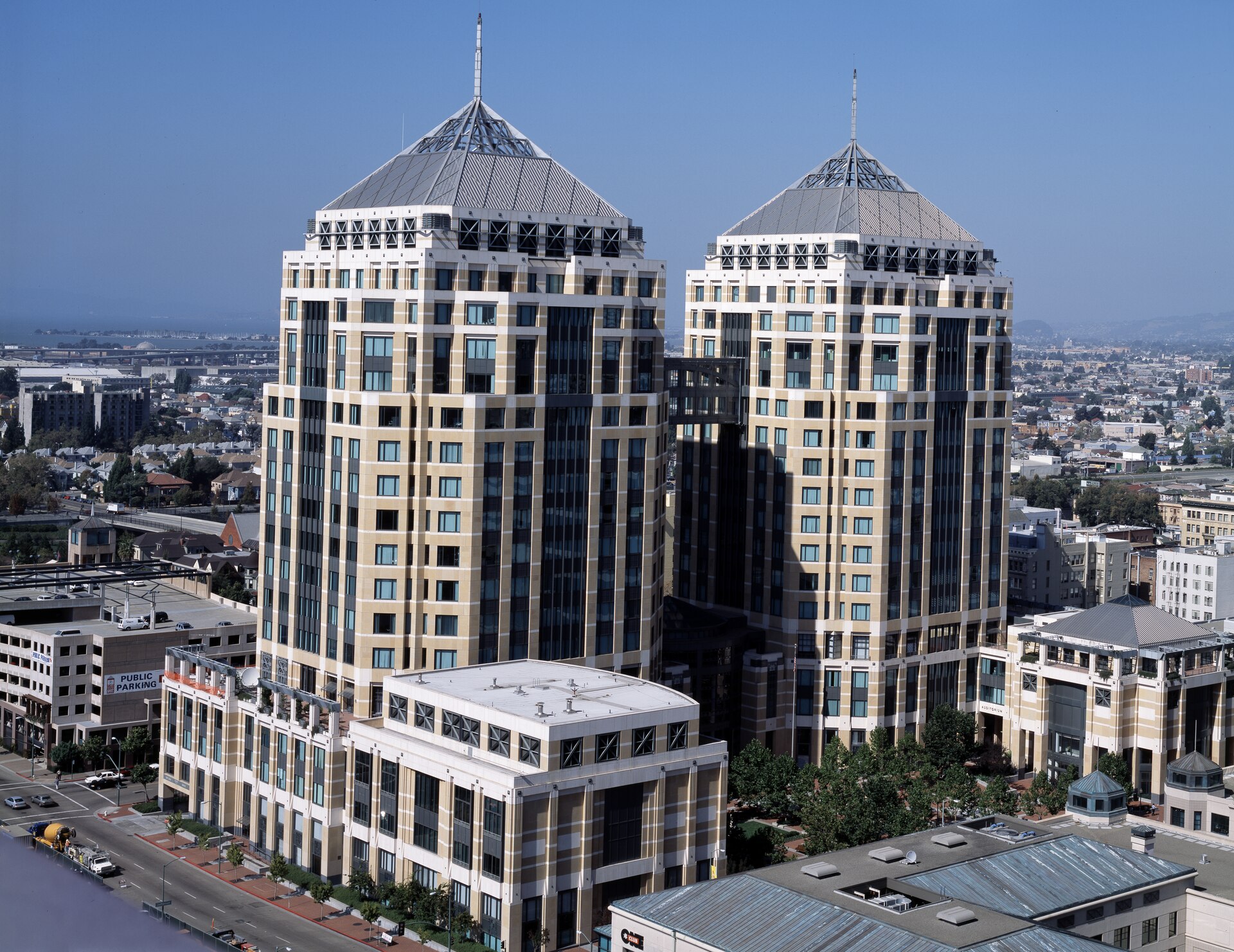 Twin federal office towers with pyramid roofs rising above downtown Oakland streets, low-rise blocks and the bay in the distance