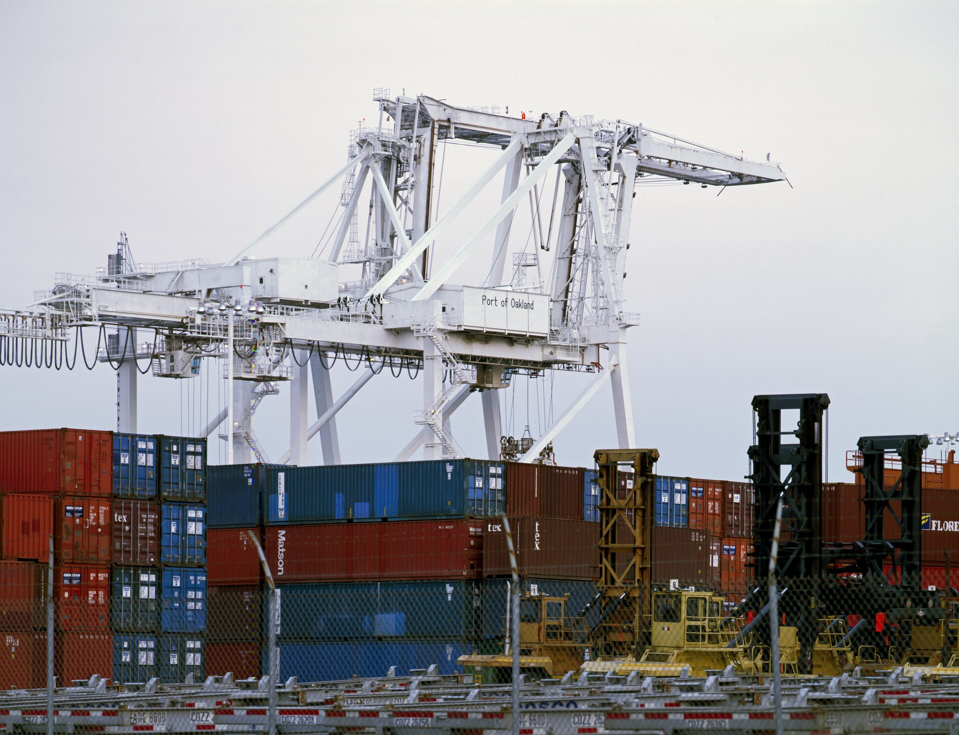 A white gantry crane lettered Port of Oakland standing over stacked shipping containers and yard equipment behind a chain-link fence