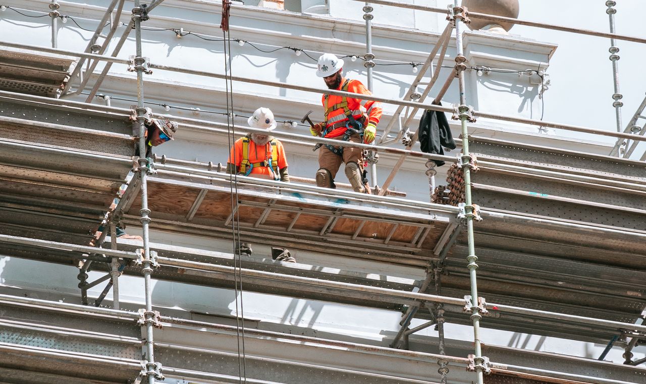 Two workers in hard hats and high-visibility gear on multi-level scaffolding against a white building facade, the retrofit and rehab work San Francisco contractors code job by job