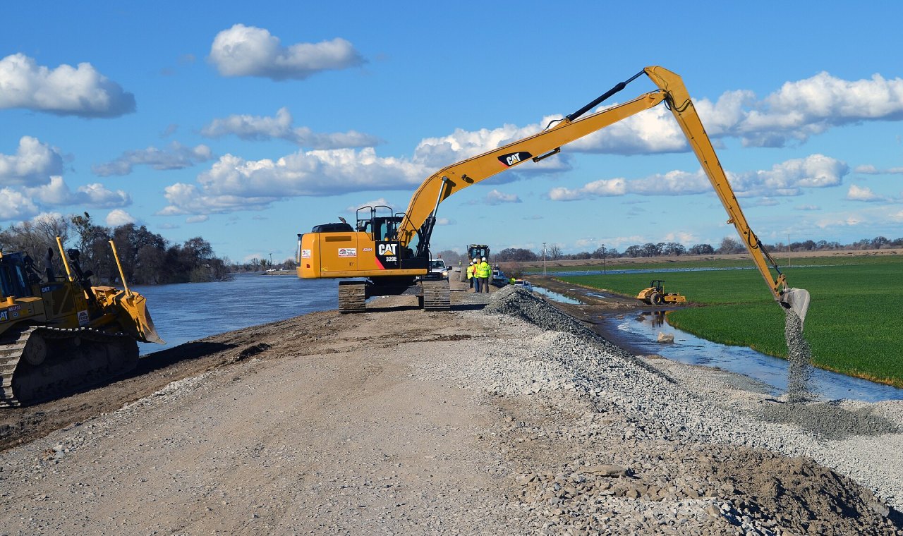 A long-reach excavator placing rock on a San Joaquin River levee bank while a dozer and crew work the crown, the heavy civil work Stockton contractors job cost