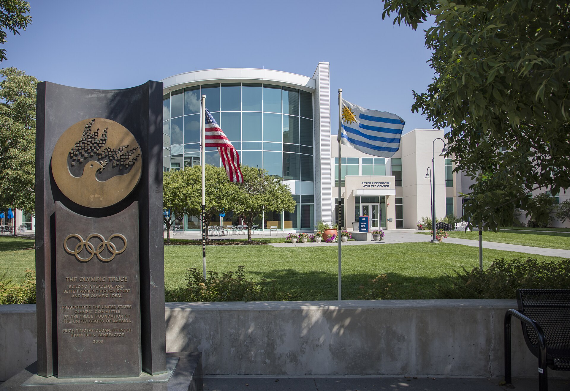 The Peter Ueberroth Athlete Center at the U.S. Olympic Training Center in Colorado Springs, a curved glass facade behind flagpoles and an Olympic Truce monument