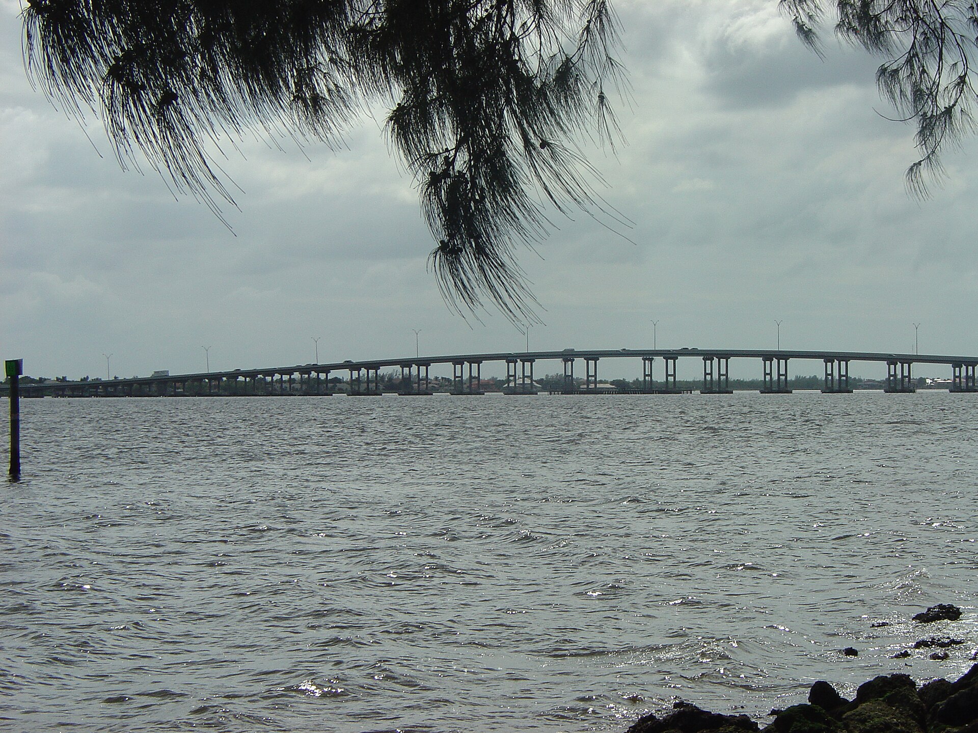 The Cape Coral Bridge crossing the Caloosahatchee River under an overcast sky, seen from the shoreline with low waterfront rooflines on the far bank