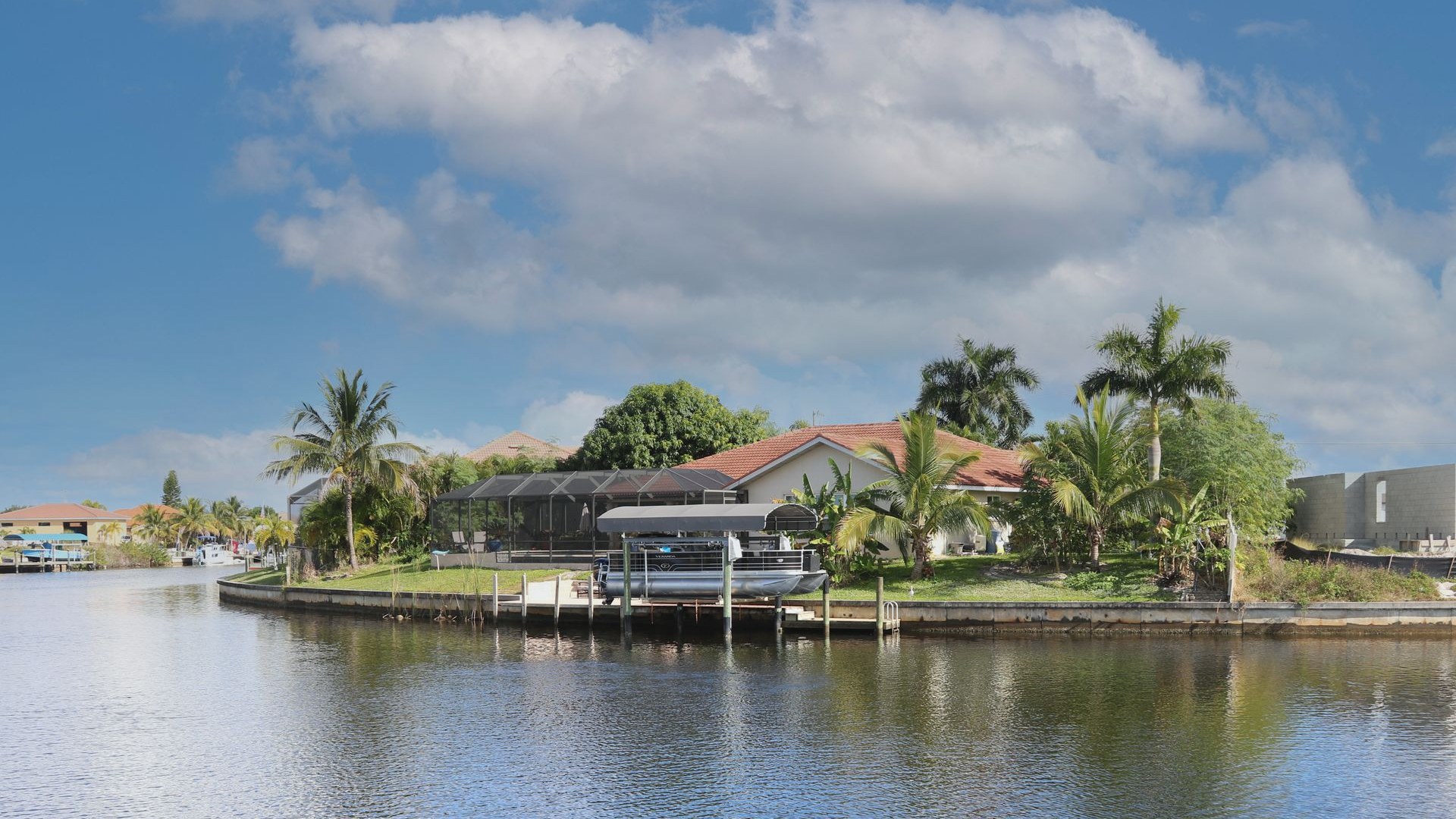 A tile-roofed Cape Coral house on a corner lot of the finger canals, screened lanai and pontoon boat on a lift at the seawall, with a concrete block house going up next door