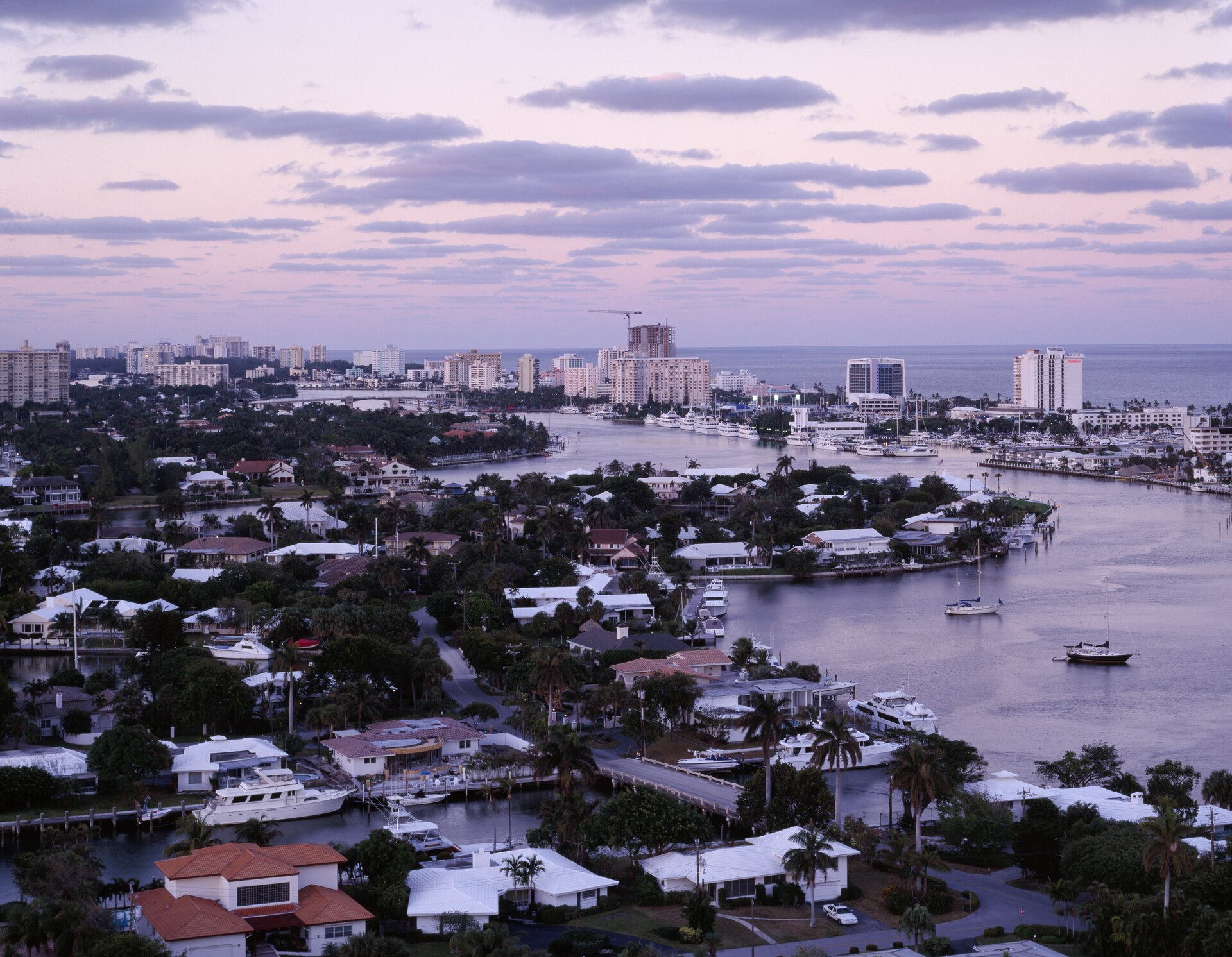 Dusk aerial over Fort Lauderdale canals lined with low-rise waterfront houses, private docks and moored boats, beach towers and a tower crane beyond