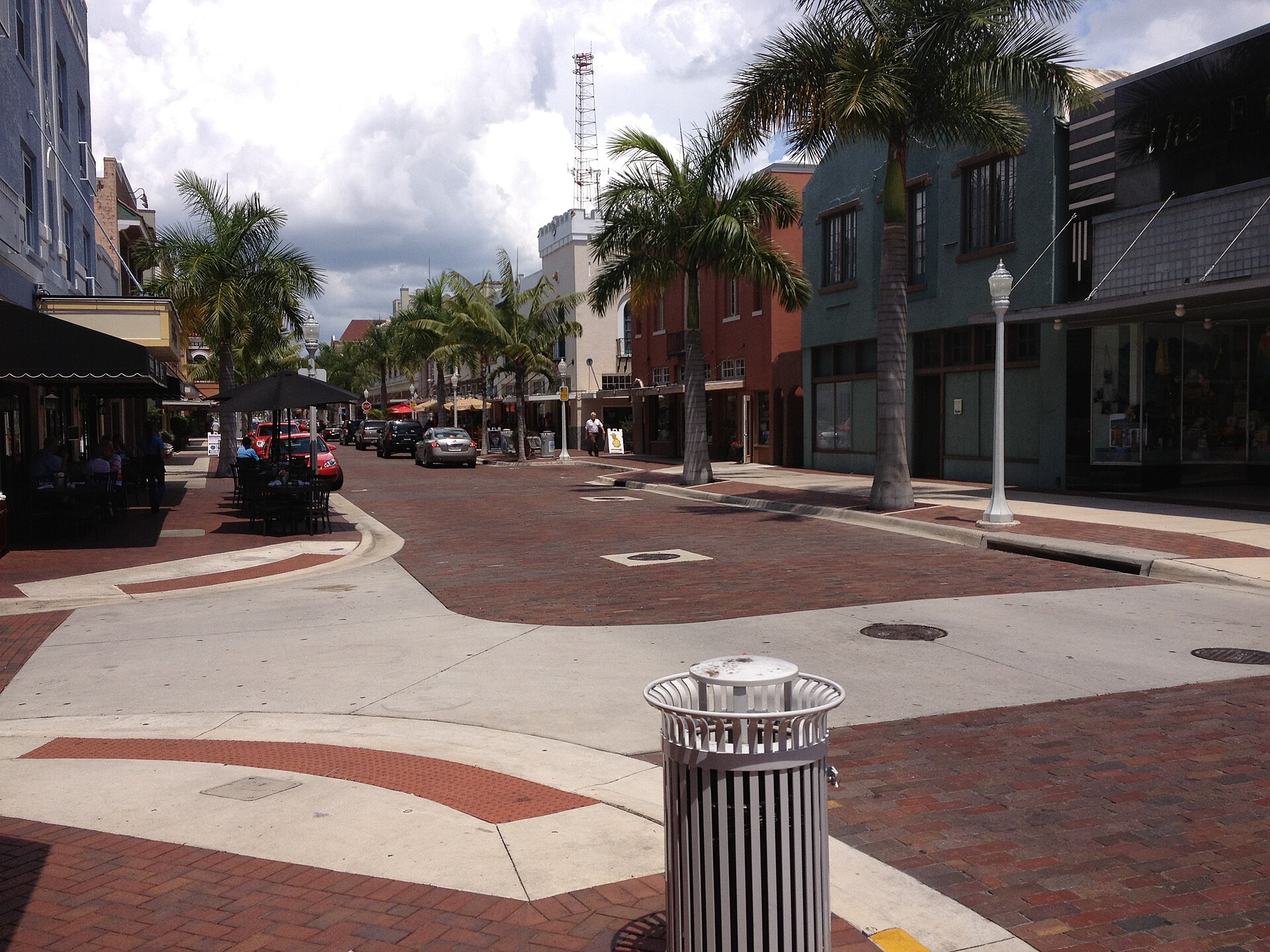 First Street in the downtown Fort Myers river district, brick paving and royal palms between painted two-story storefronts with a sidewalk cafe
