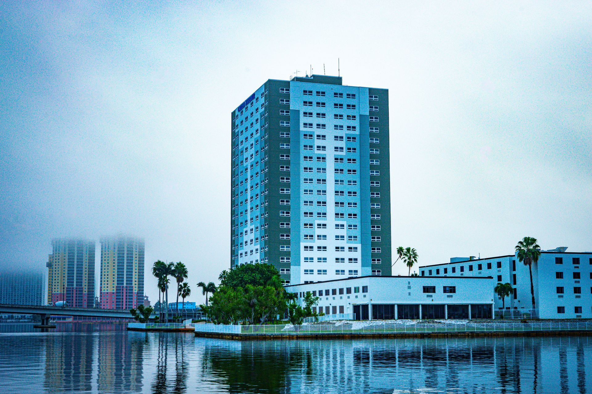 A tall white and grey residential tower on the Caloosahatchee riverfront in Fort Myers under an overcast sky, low bridge and pastel mid-rise blocks across the water