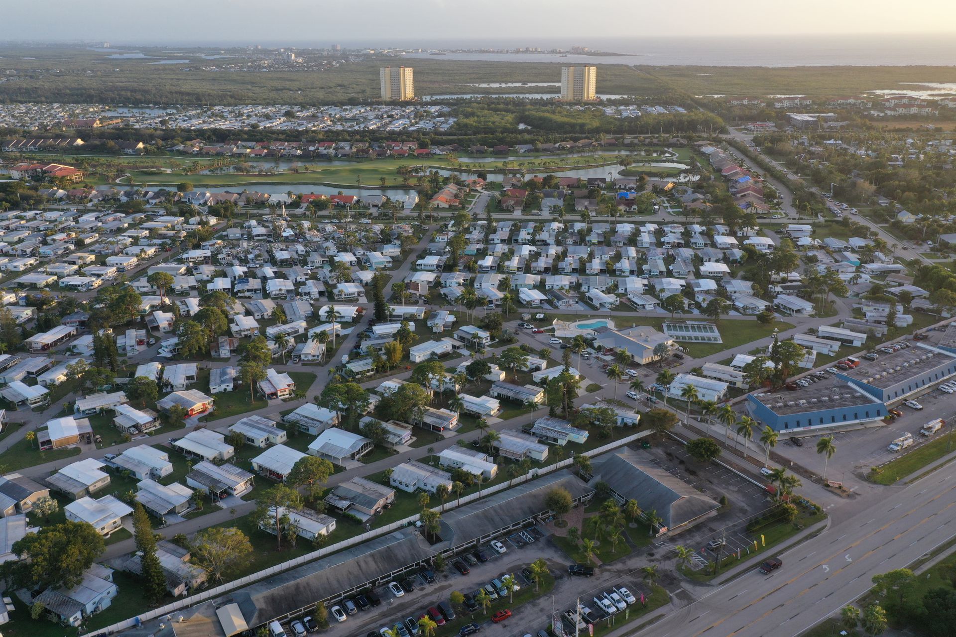 Evening aerial over Fort Myers looking toward the coast, rows of single-story manufactured homes and tile-roofed houses around a golf course, two condominium towers and the Gulf on the horizon