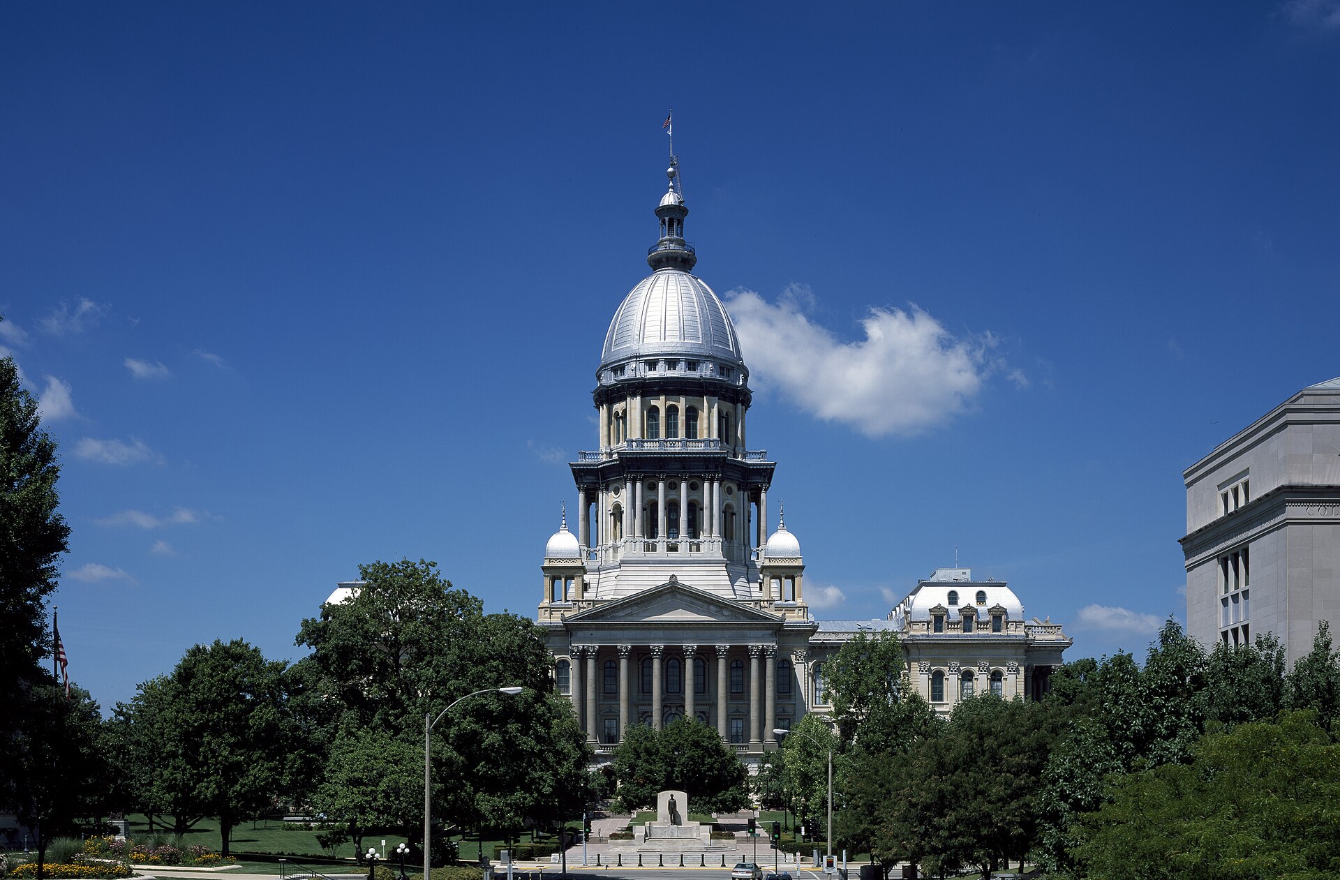The Illinois State Capitol in Springfield under a deep blue summer sky, its silver ribbed dome and columned portico rising above the trees on the capitol grounds