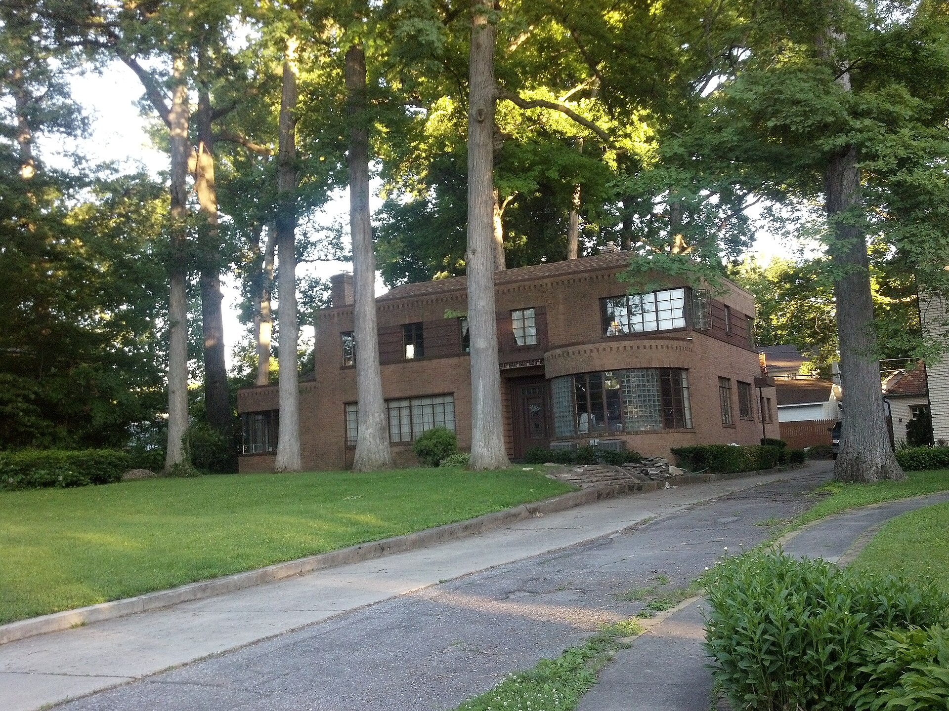A 1930s brown brick house with rounded corner windows on a shaded Fort Wayne residential street, mature trees and a cracked concrete driveway