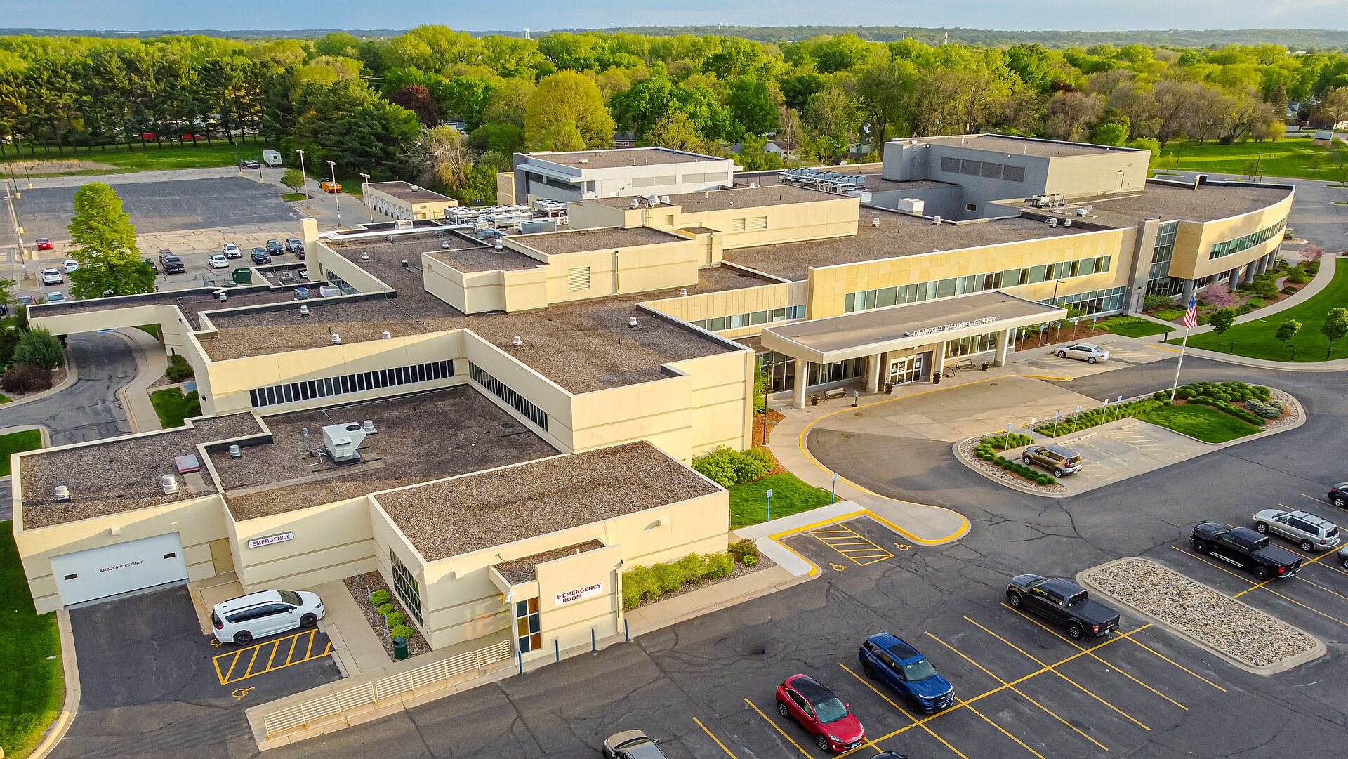Aerial view of the Olmsted Medical Center campus in Rochester, Minnesota, its low clinic wings, emergency entrance canopy and parking lots ringed by spring trees