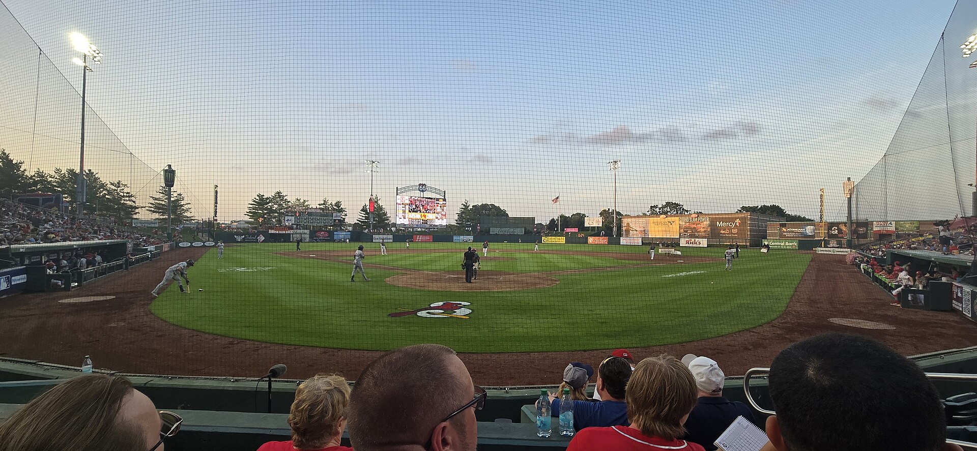 Route 66 Stadium in downtown Springfield, Missouri at dusk, a Springfield Cardinals game under way with the infield and outfield boards lit and the crowd filling the stands