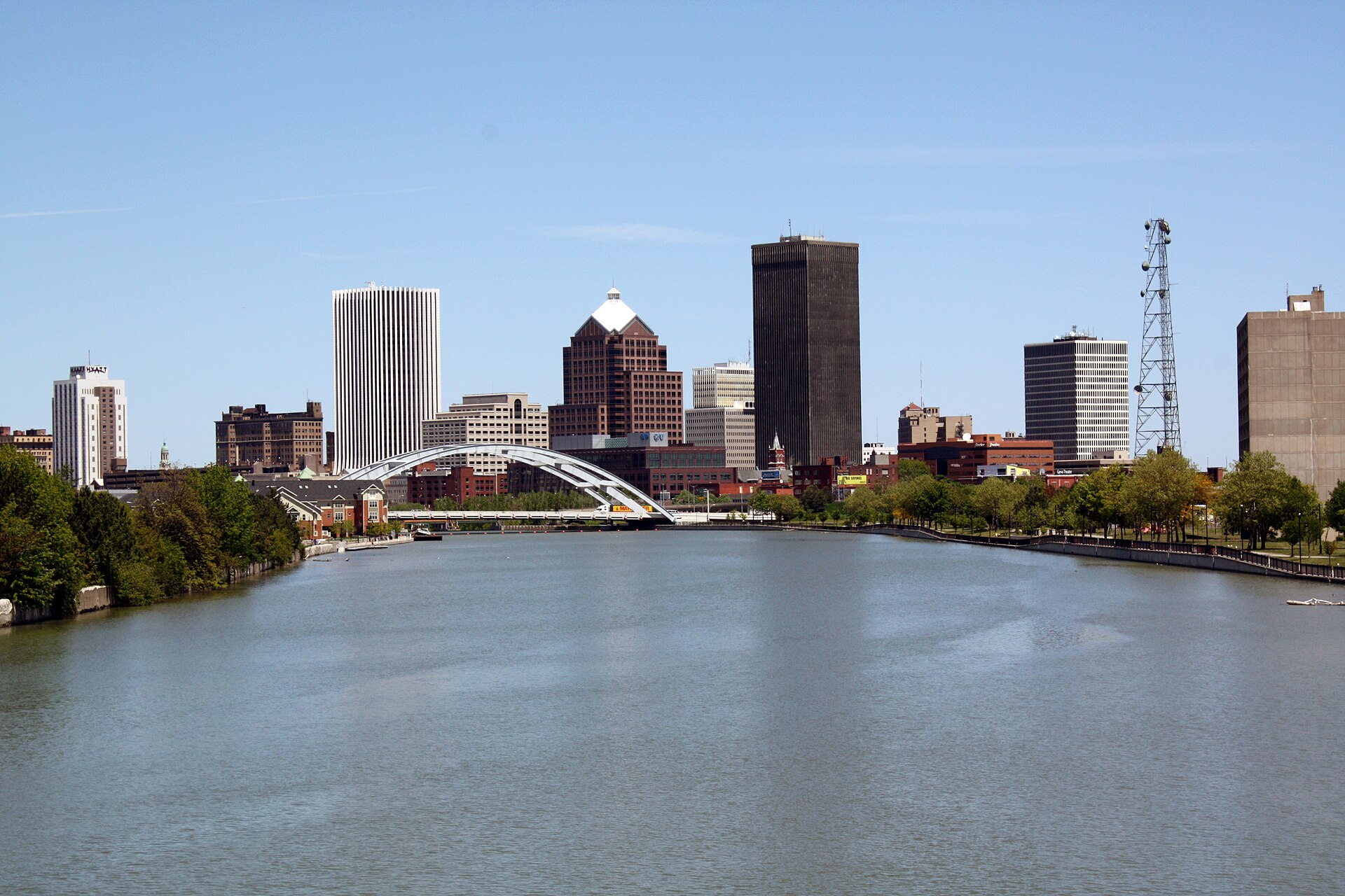 The downtown Rochester, New York skyline seen up the Genesee River, office towers behind a white arched road bridge and tree-lined riverbanks