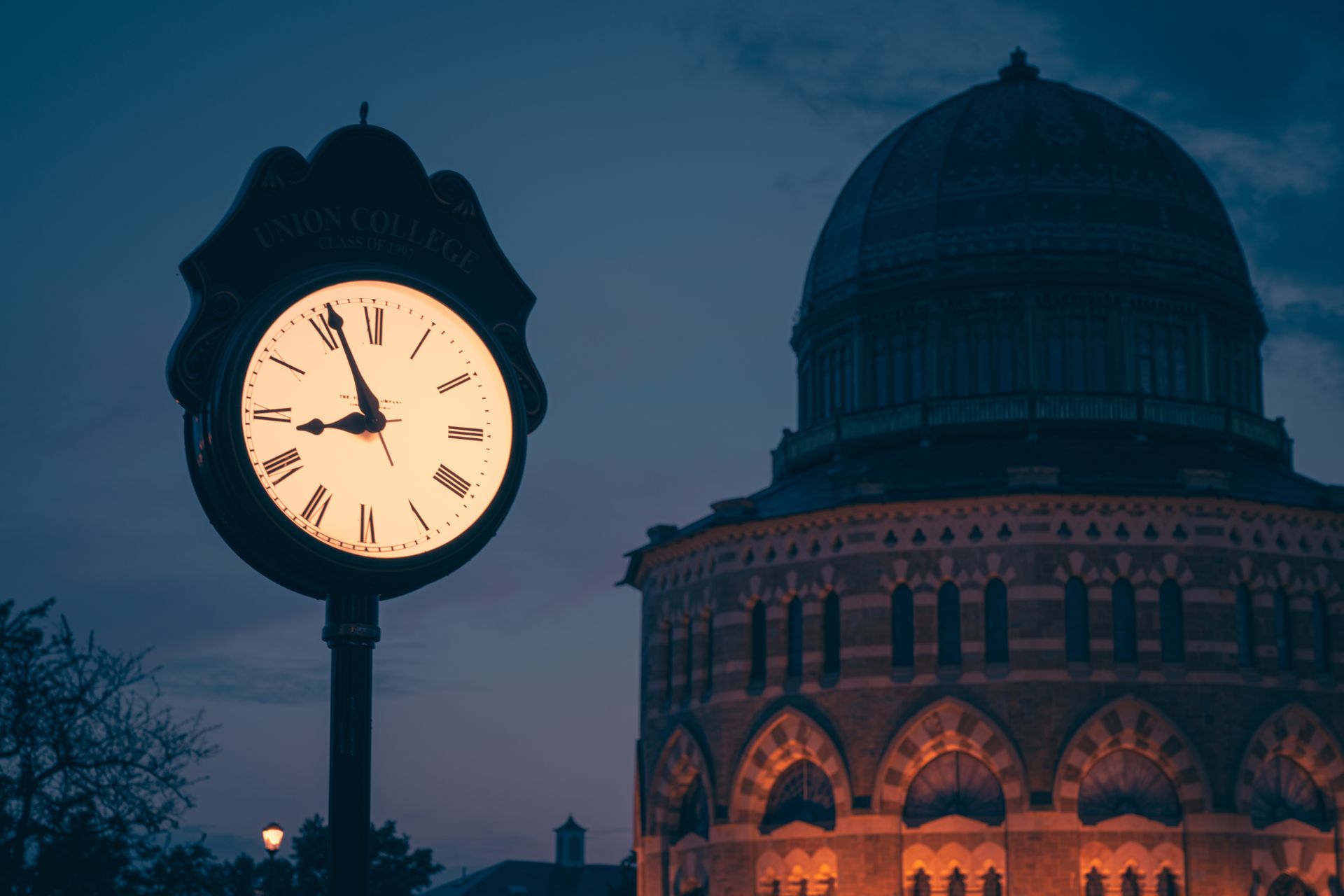 The Union College clock in Schenectady at dusk, its lit face in front of the sixteen sided Nott Memorial and its ribbed dome