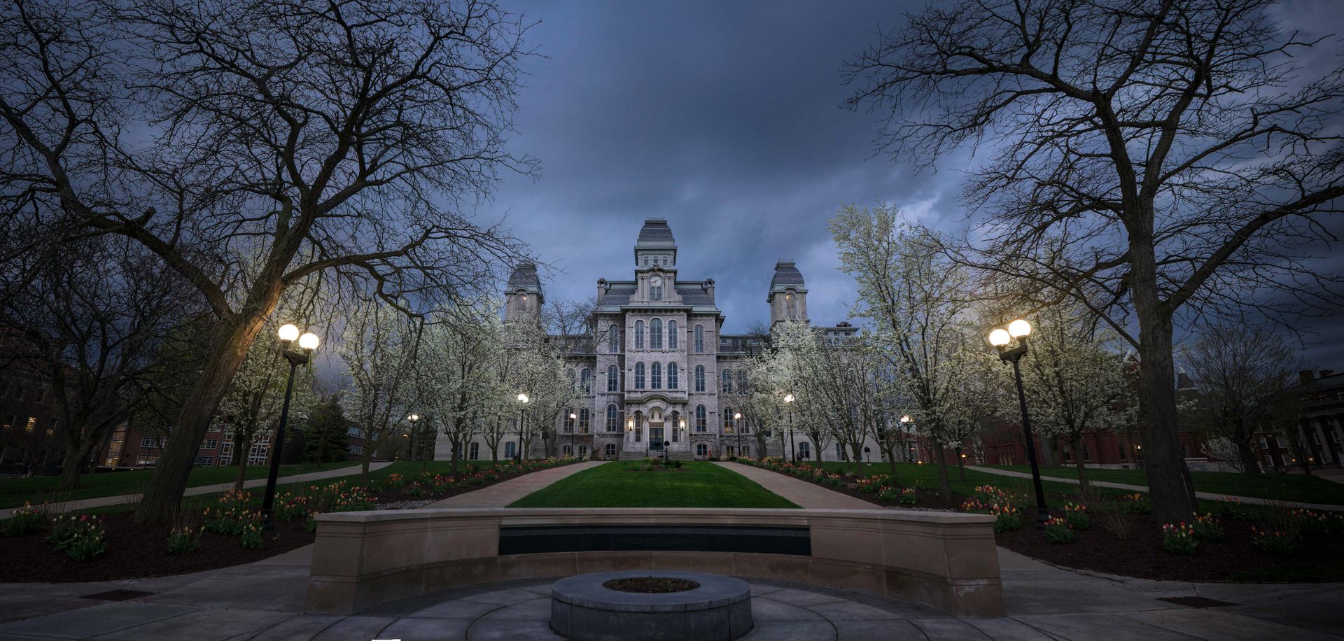 The Hall of Languages at Syracuse University lit at dusk, a grey stone building with towers and a clock, flowering trees and tulip beds on the quad in front of it