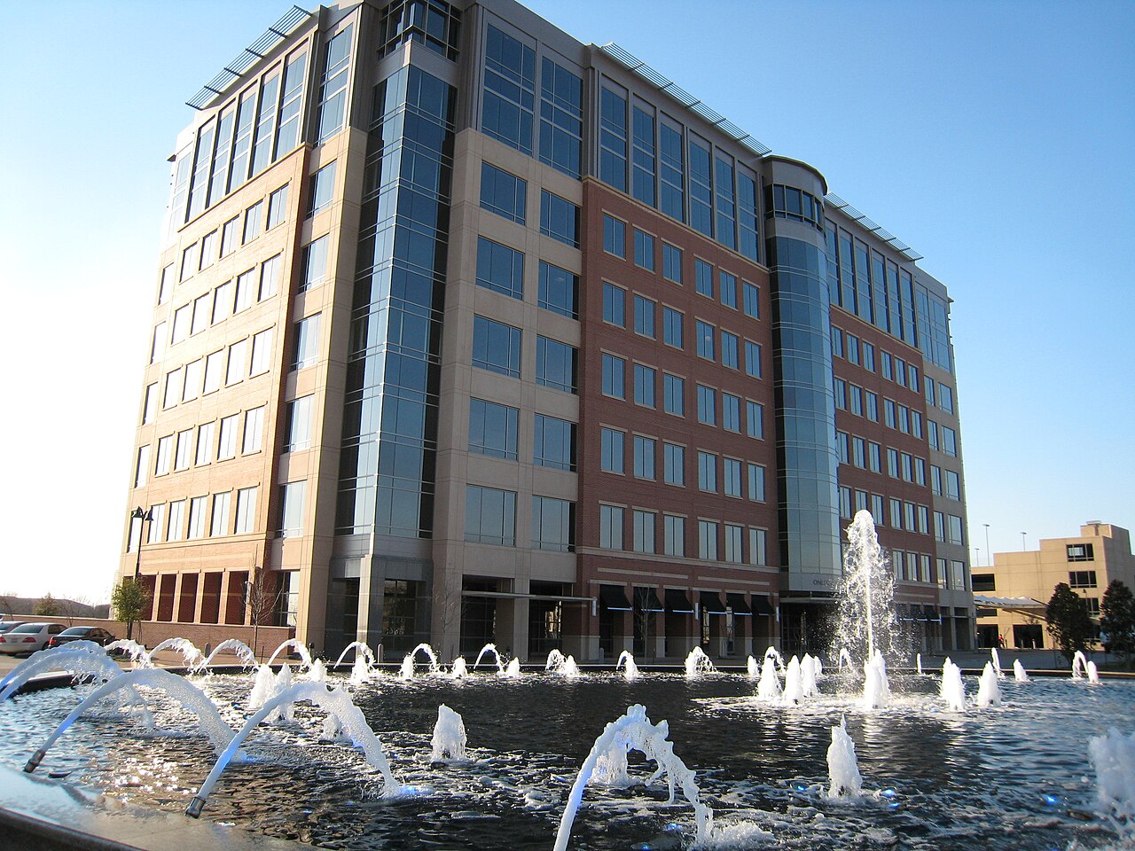 A mid-rise brick and glass corporate office building behind a fountain plaza in Plano, the headquarters fit-out market FinTruction supports with WIP reporting