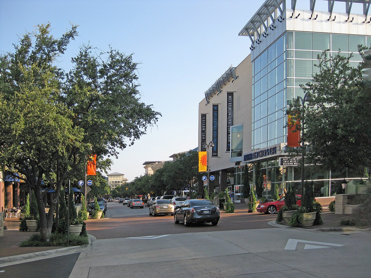 A tree-lined street of ground-floor shops and restaurants below a glass office building in Plano, Texas, the suburban commercial market FinTruction job costs