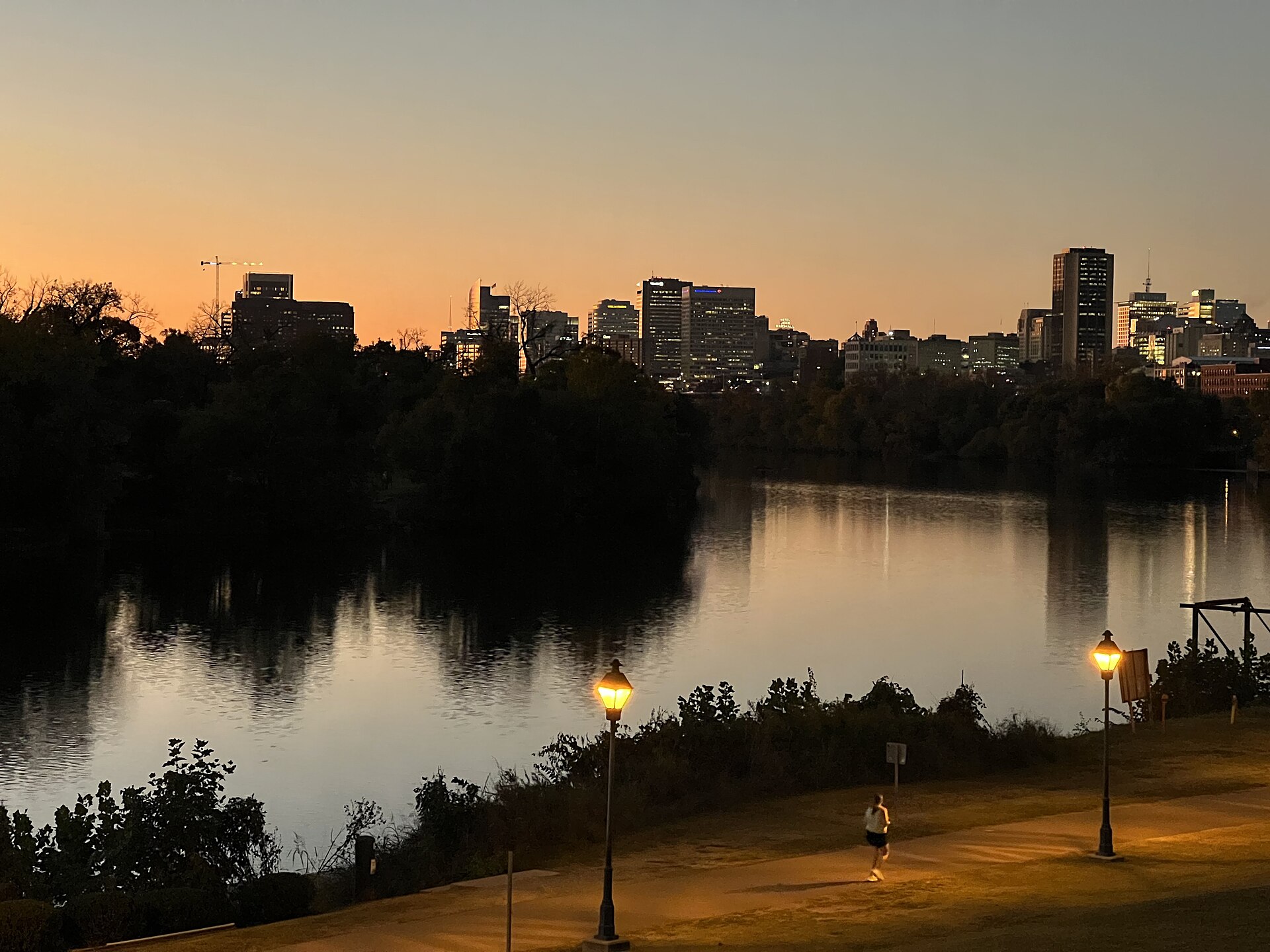 Richmond skyline at sunset seen across the James River from Shockoe Bottom, a tower crane at the left edge and lamp posts along the riverside path