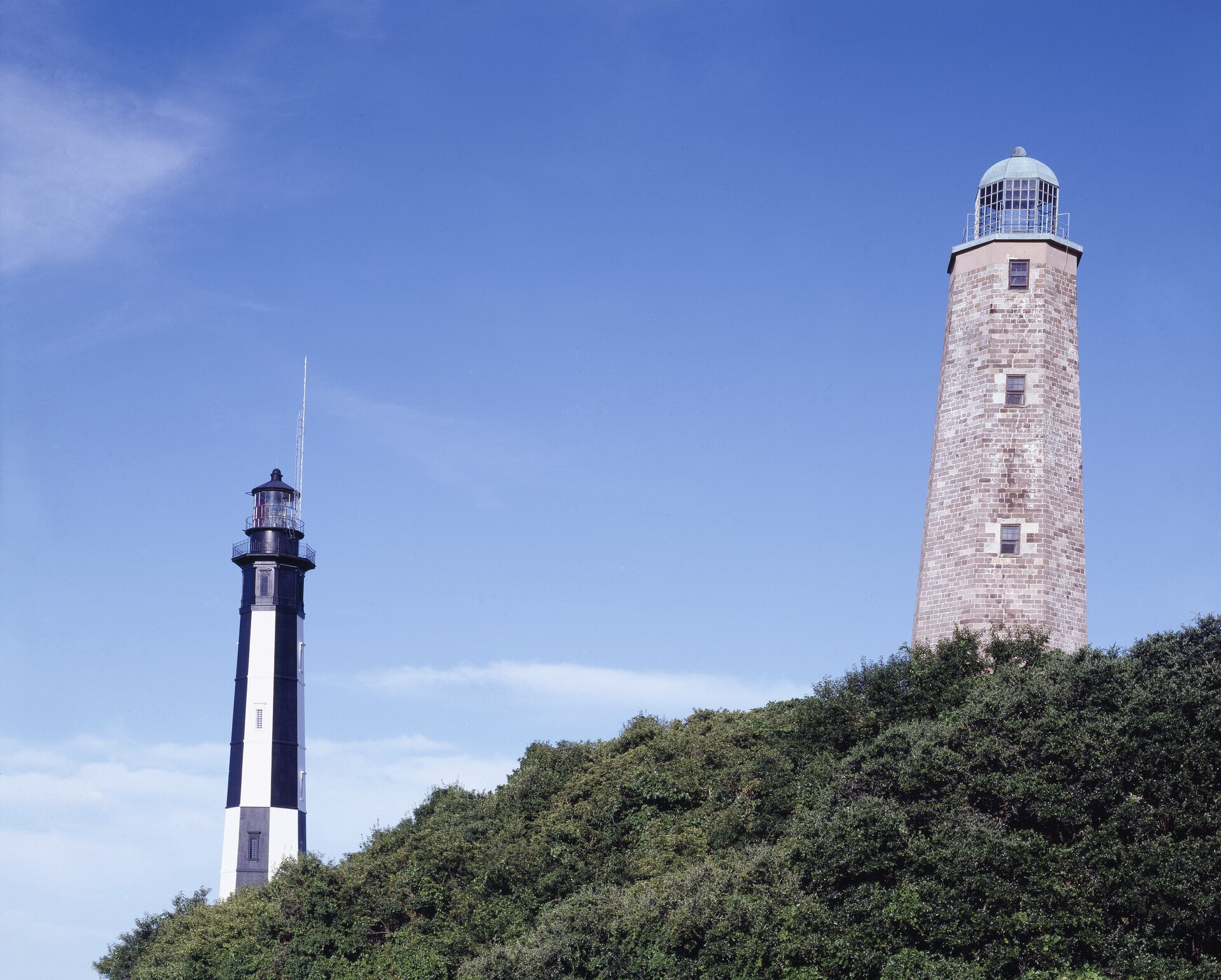 The old sandstone Cape Henry Lighthouse and the taller black and white cast iron tower beside it, rising from wooded dunes in Virginia Beach