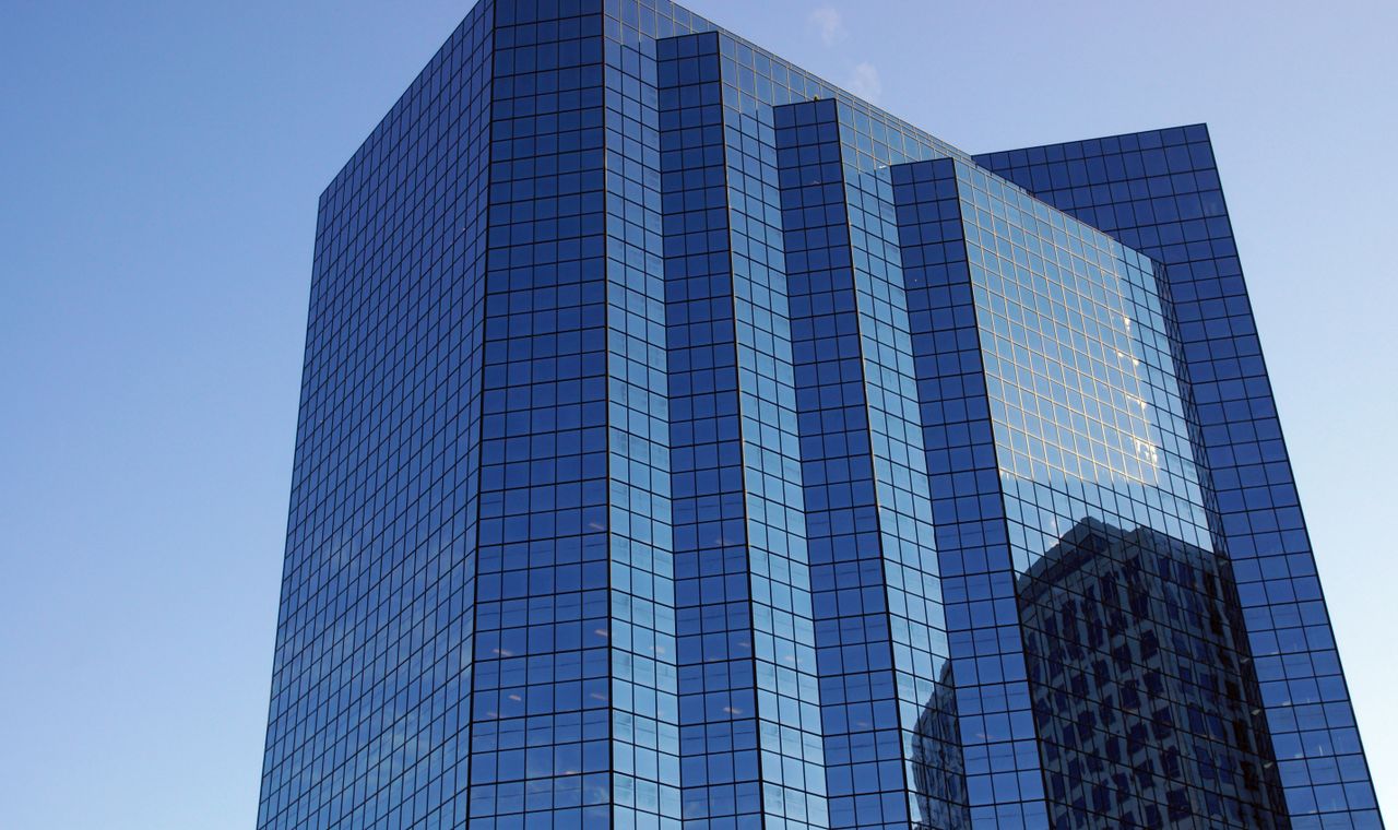 A blue glass office tower photographed from street level with a neighboring high-rise mirrored in its facade, the downtown Bellevue tower work FinTruction job costs